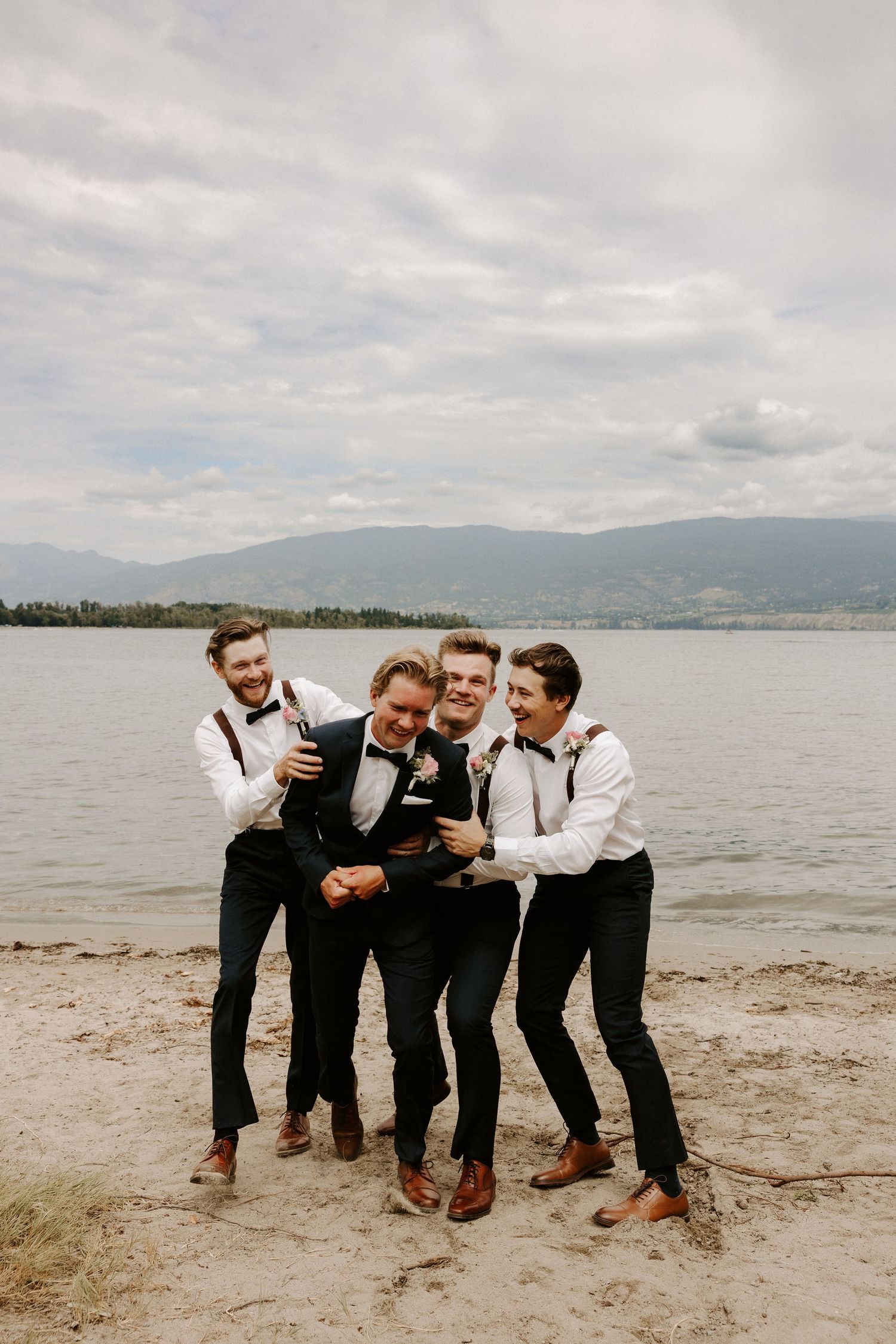 Groomsmen in matching black and white attire share laughs on scenic beach with mountain views.
