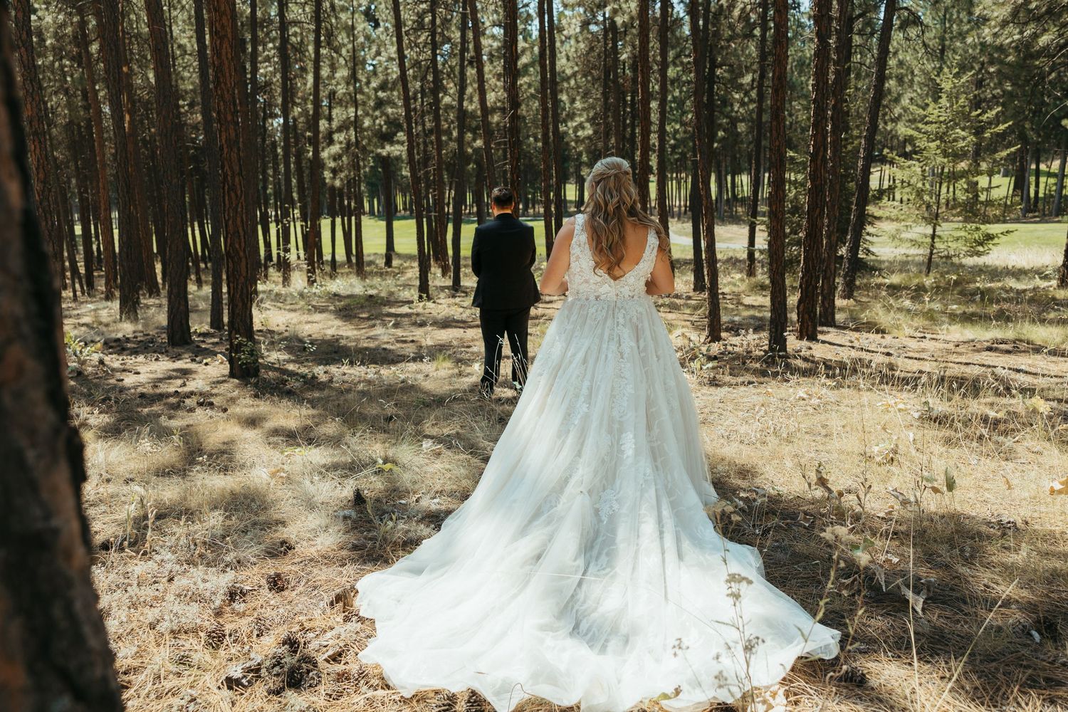 Bride in flowing white dress follows groom through sunlit pine forest.
