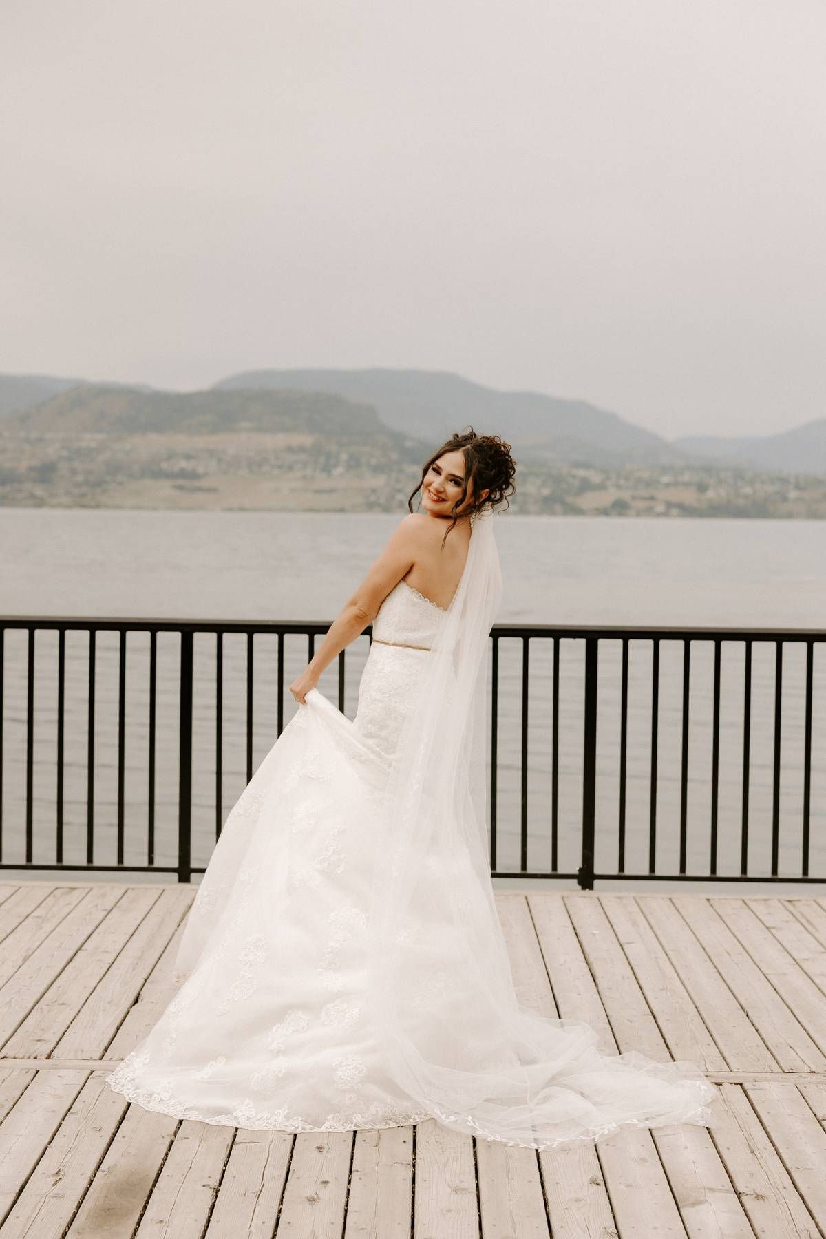 A bride in a flowing white wedding gown poses gracefully on a deck overlooking a misty lake and mountains.