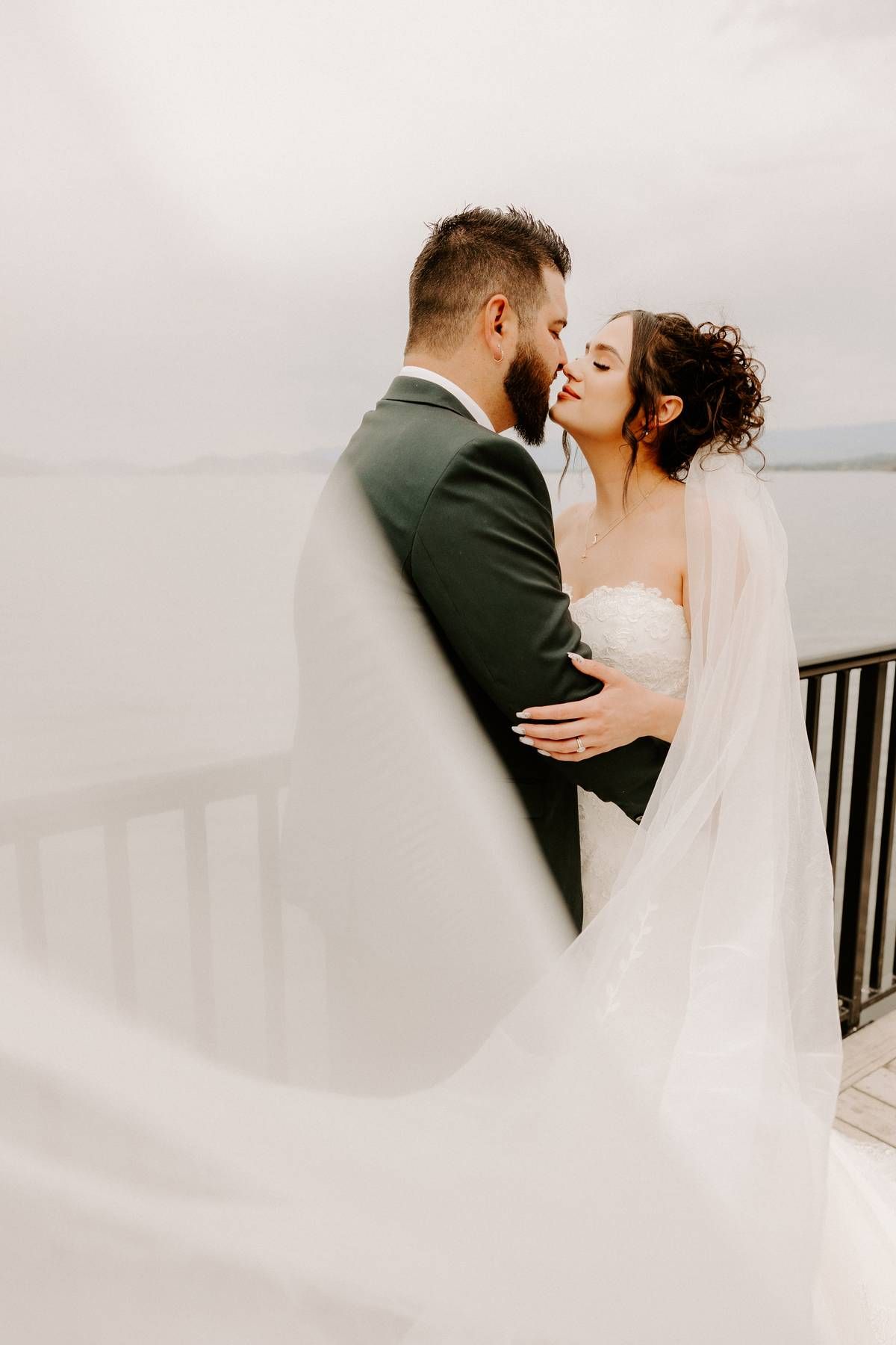 Romantic wedding couple embraces on a balcony as bride's veil flows in the wind during an intimate moment.