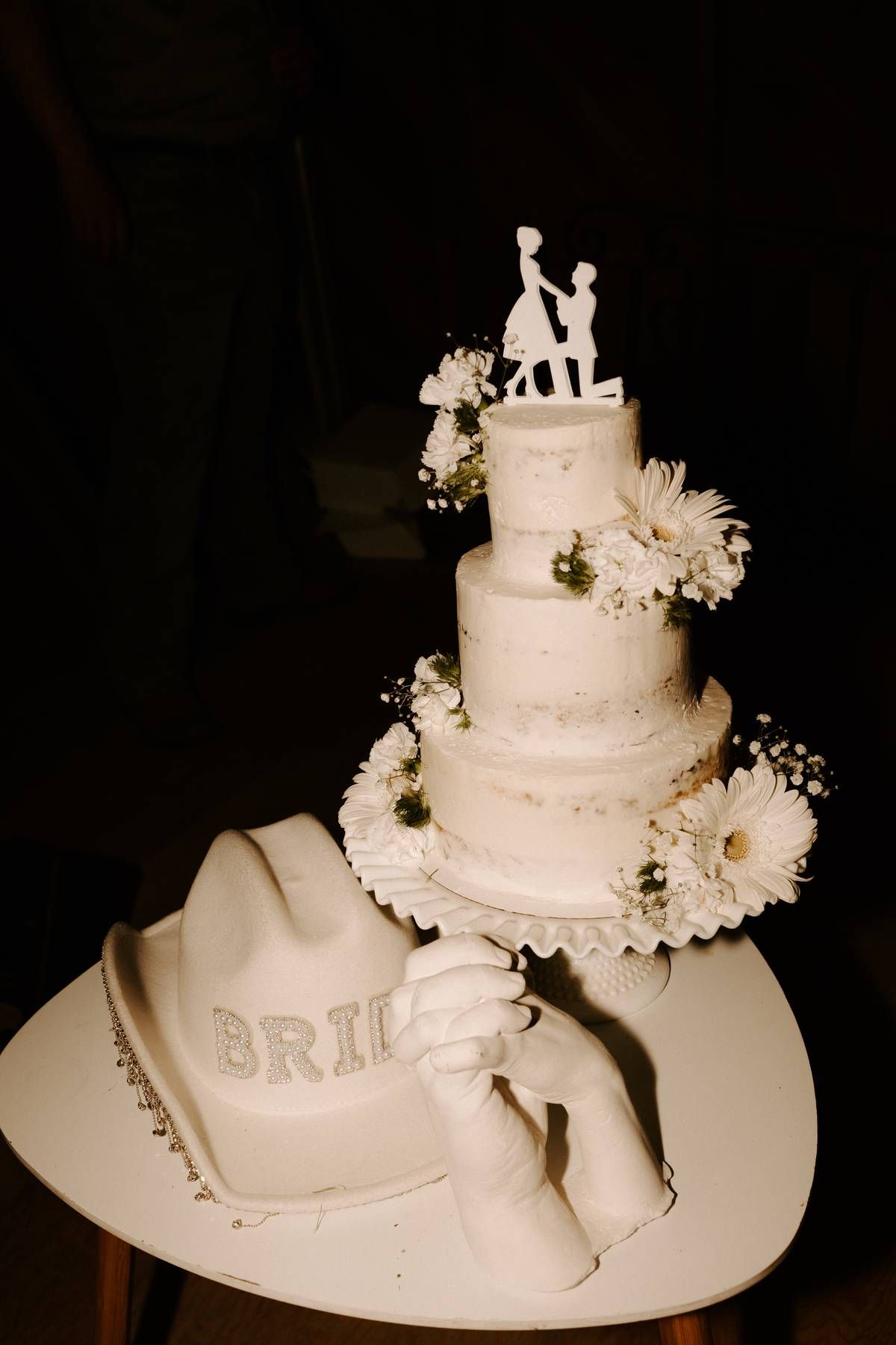 Elegant white tiered wedding cake decorated with daisies and a proposal topper, accompanied by a decorative bride's cowboy hat.