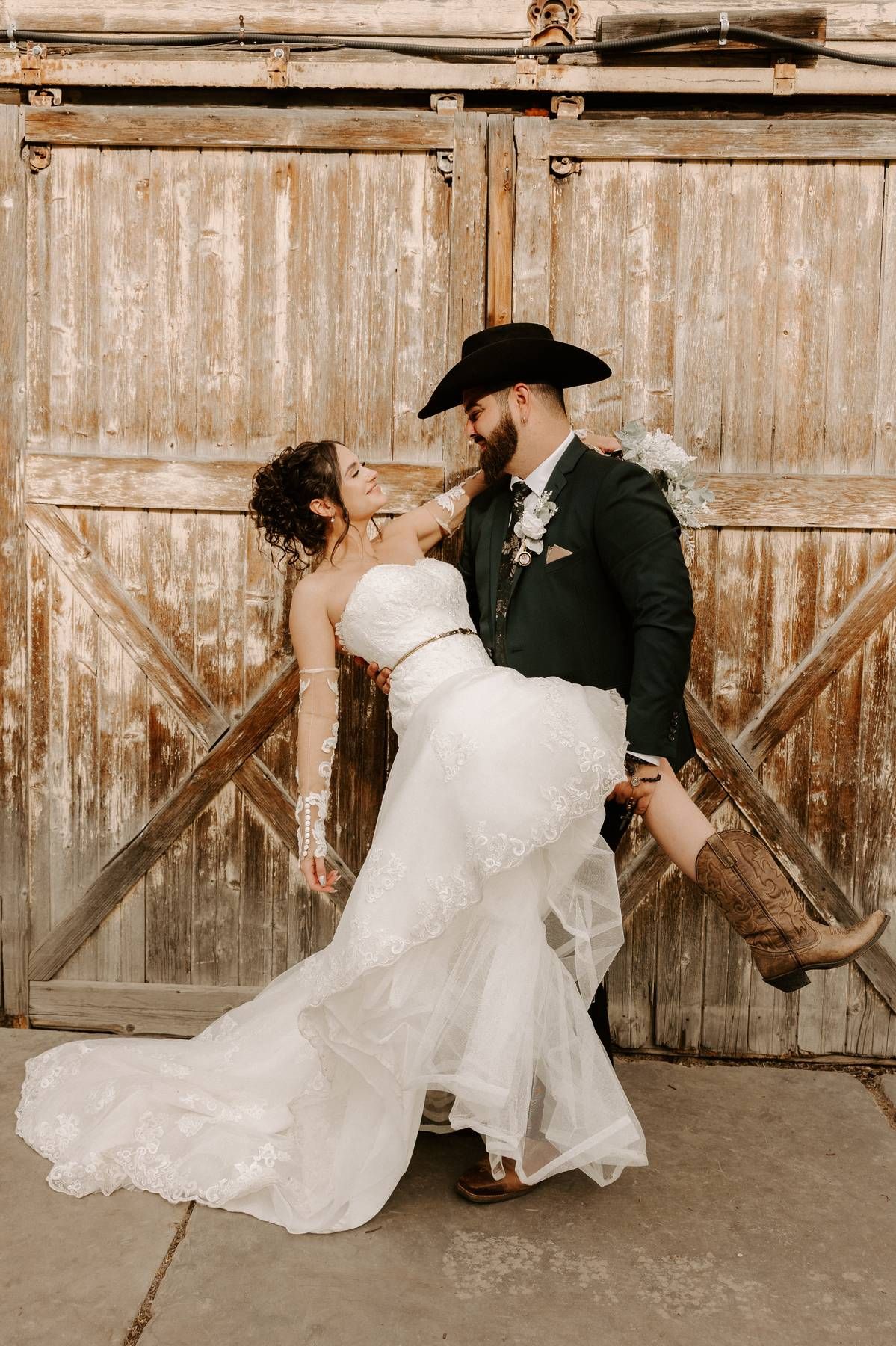 A couple in western wedding attire poses against rustic barn doors in a romantic dip pose.