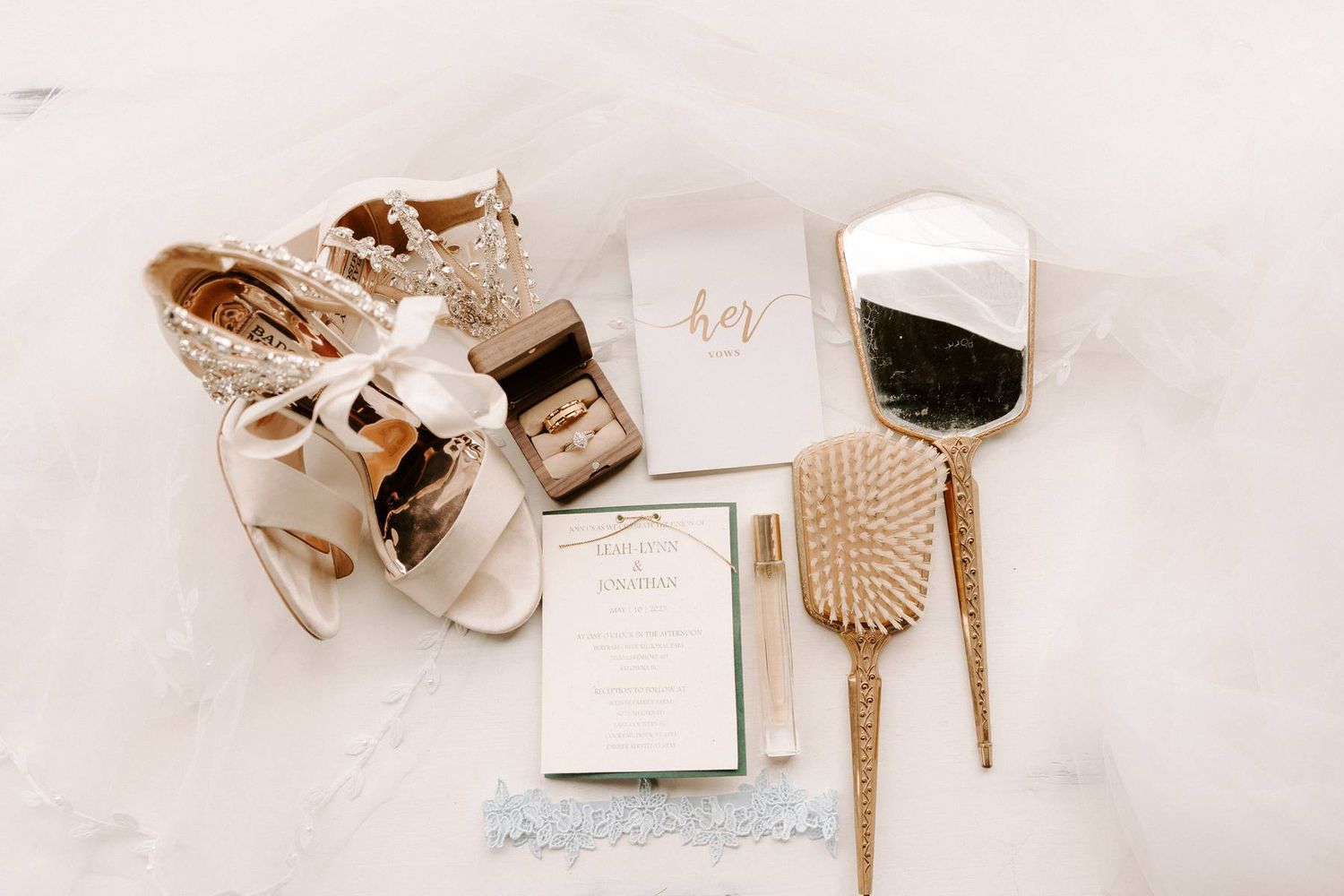 Wedding accessories including gold heels, hair brush, and stationery arranged on white fabric for a bridal flat lay.