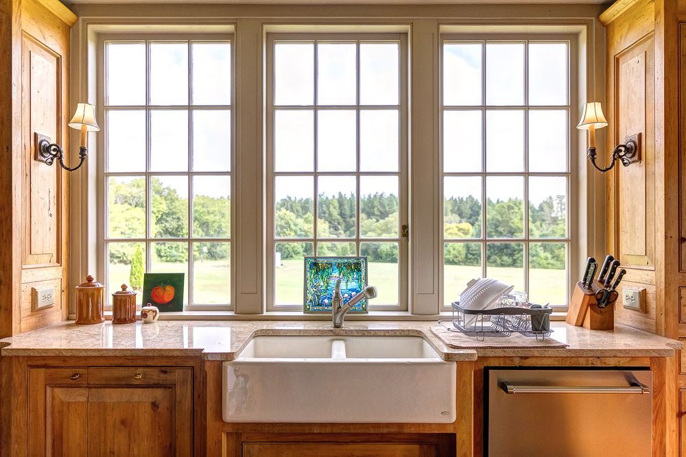 Farmhouse sink below three large windows with natural light and wooden cabinetry in a traditional kitchen.