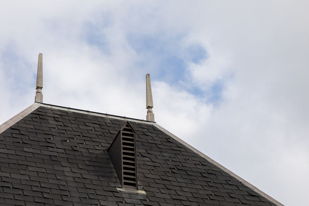 Black slate roof with twin chimneys against blue cloudy sky.