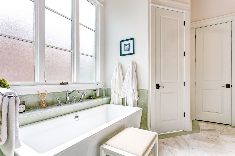 Bright modern bathroom with freestanding tub, large windows, and crisp white decor.
