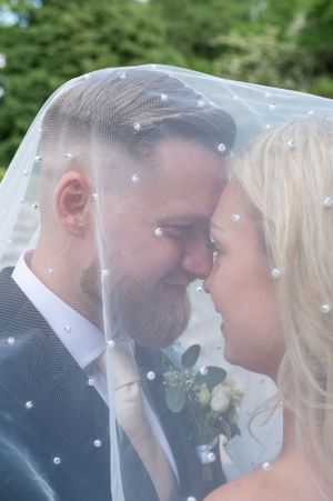 Bruden och brudgummen delar ett intimt ögonblick under en pärlprydd brudslöja i en grönskande miljö.