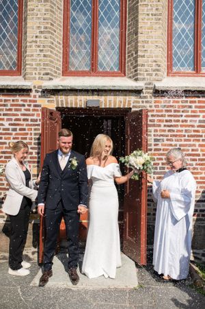 Ett nygift par kommer ut ur en tegelkyrka i ett konfettiregn, åtföljda av en präst och en bröllopsgäst, en solig dag.