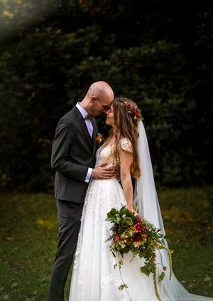 Bruden och brudgummen utbyter en mjuk kyss utomhus; bruden bär en blomsterkrans och håller i en fyllig höstbukett.