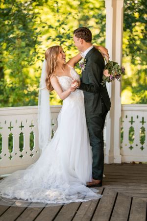 Bruden och brudgummen delar en romantisk stund på verandan till ett vitt lusthus; bruden bär en spetsklänning och håller i en lila blomsterb