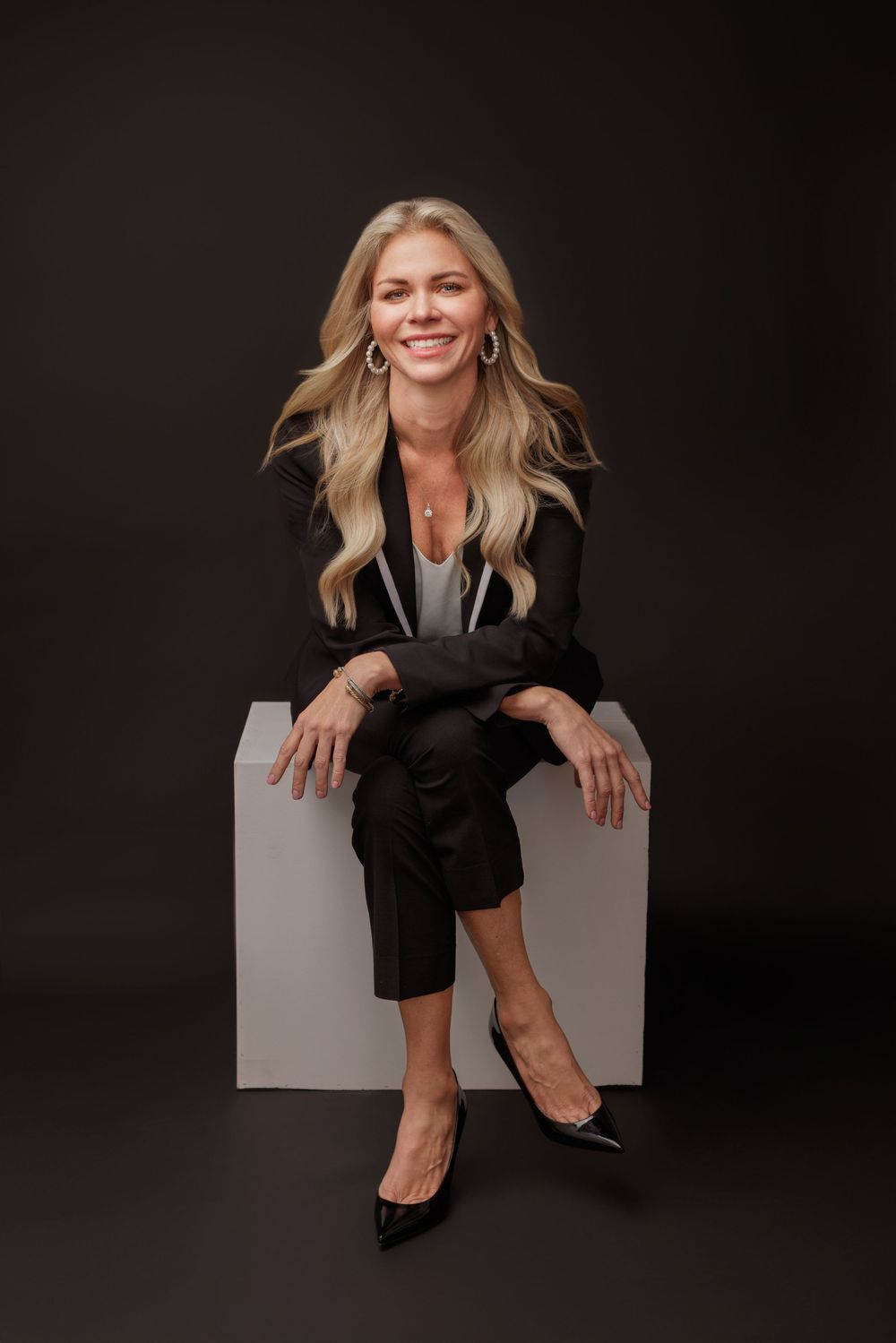 Series of studio portraits of a woman in black clothing sitting casually on a white cube against a dark backdrop.