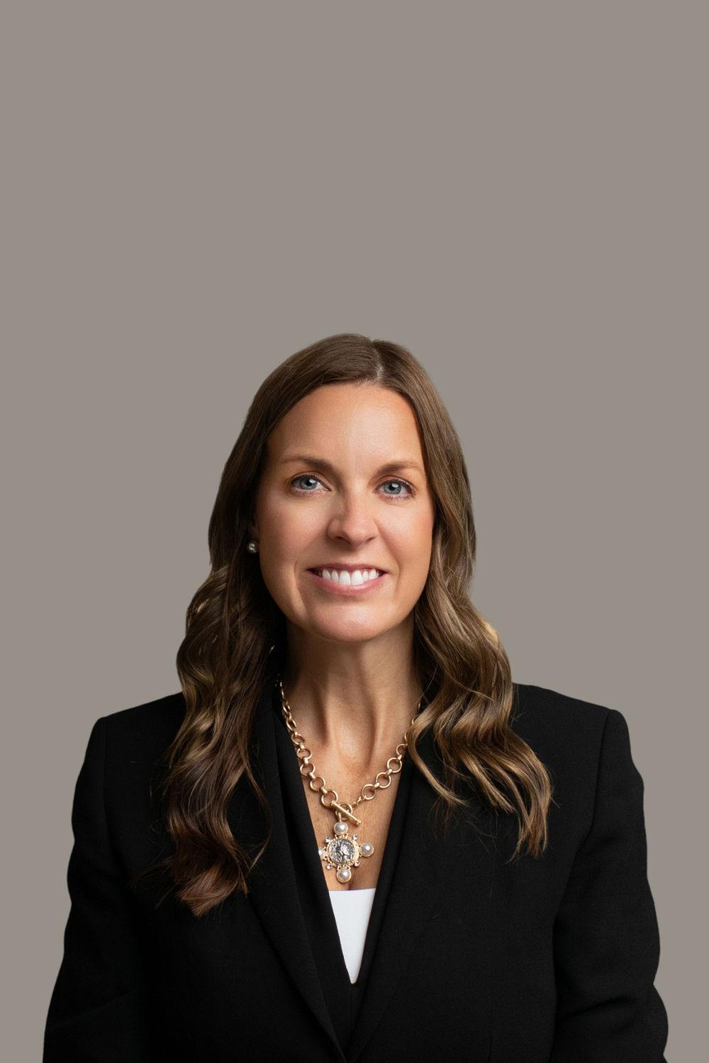 Professional headshot of a businesswoman wearing a black blazer and statement necklace against a grey background.