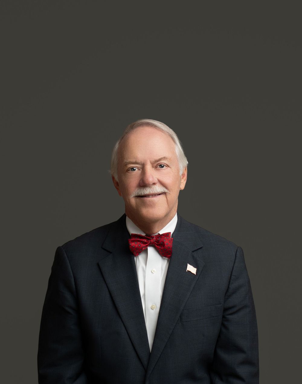 Formal portrait of person in black tuxedo and red bowtie against dark background.