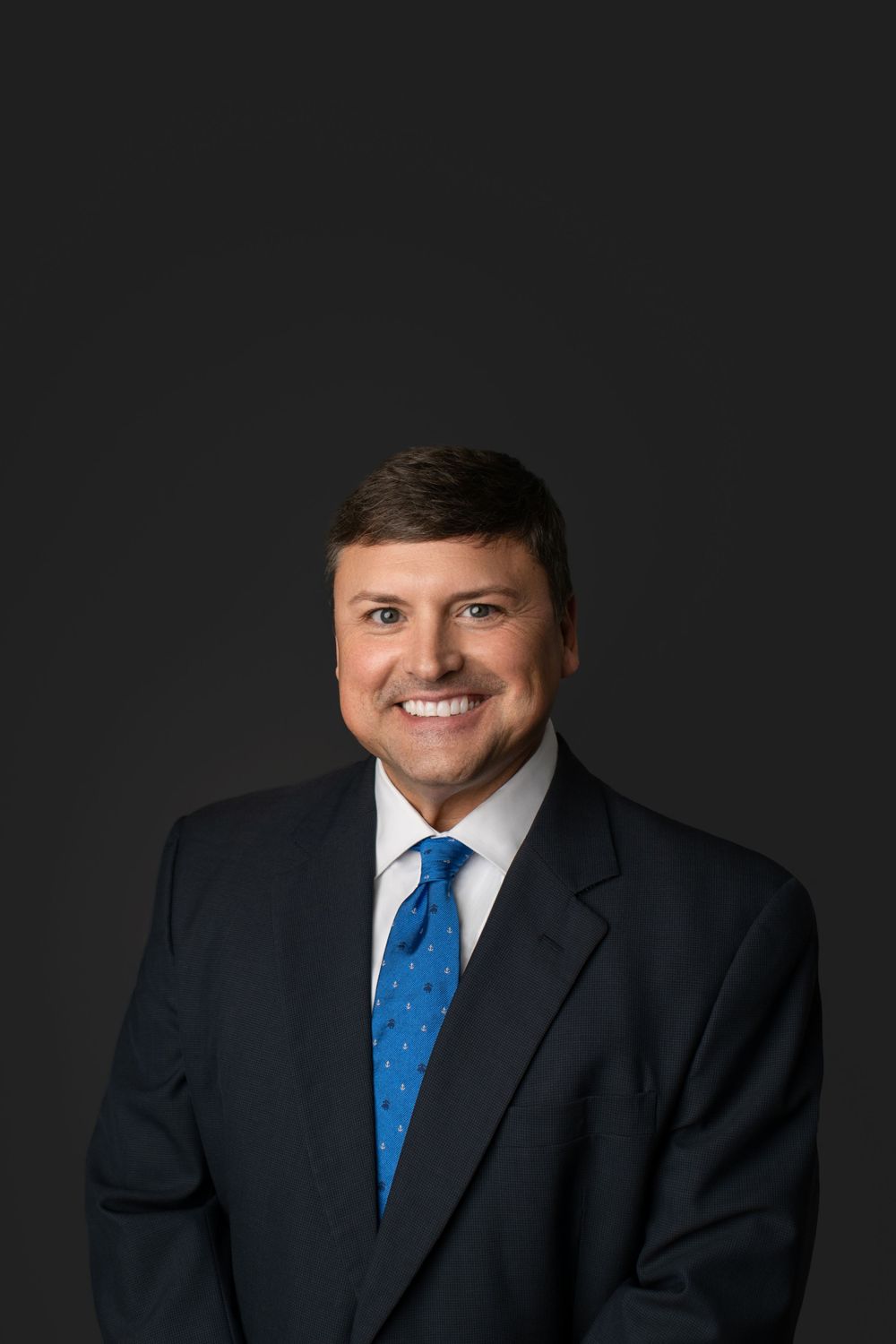 Professional headshot of individual in dark suit with blue polka dot tie against black background.