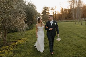 Wedding couple strolls through a sun-drenched meadow during golden hour with blooming trees in the background.