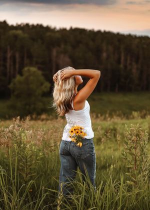Blonde woman in white tank top and jeans holding sunflowers in a lush green field at golden hour.