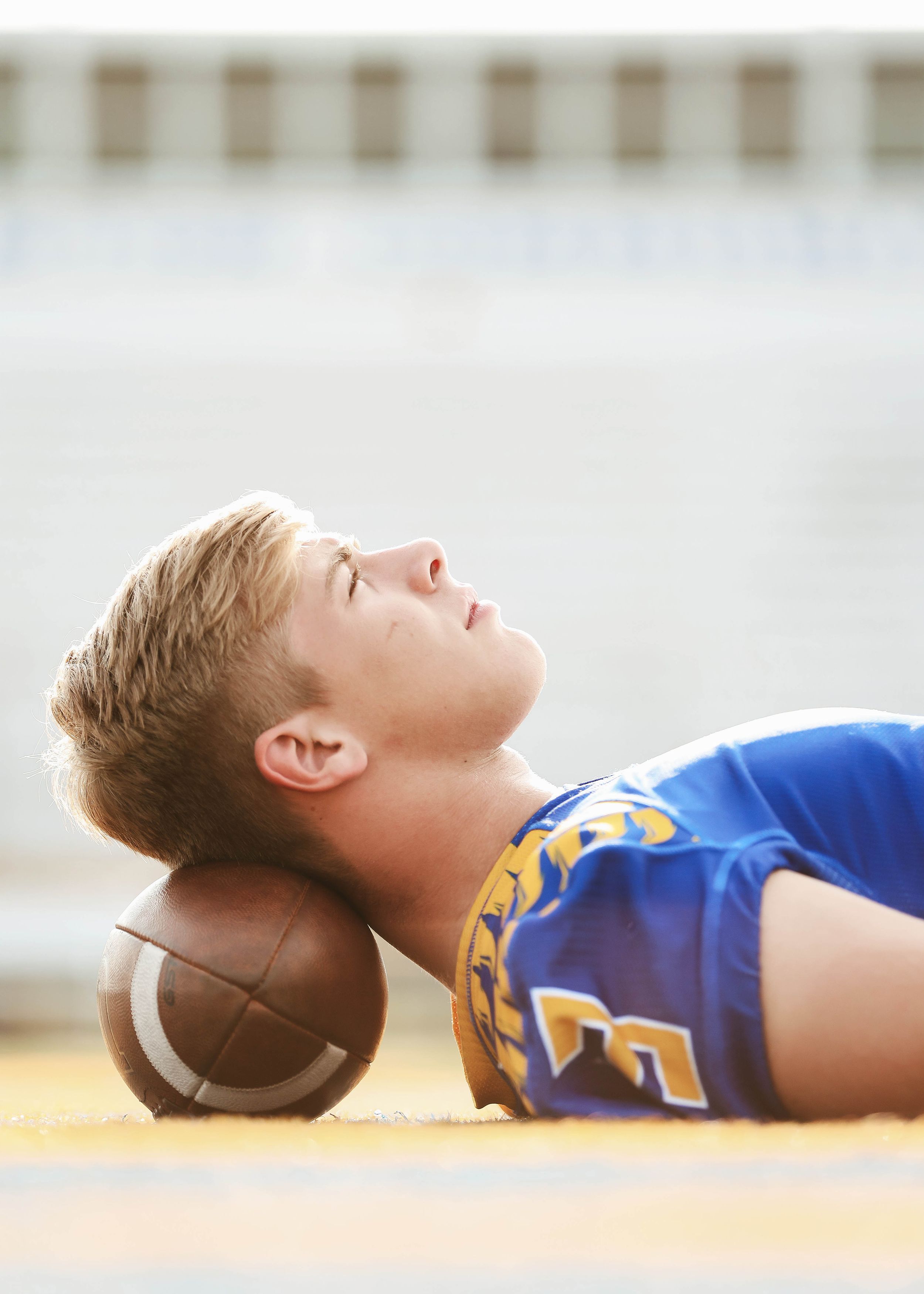 Athlete in blue uniform lying on ground with football in dramatic pose.