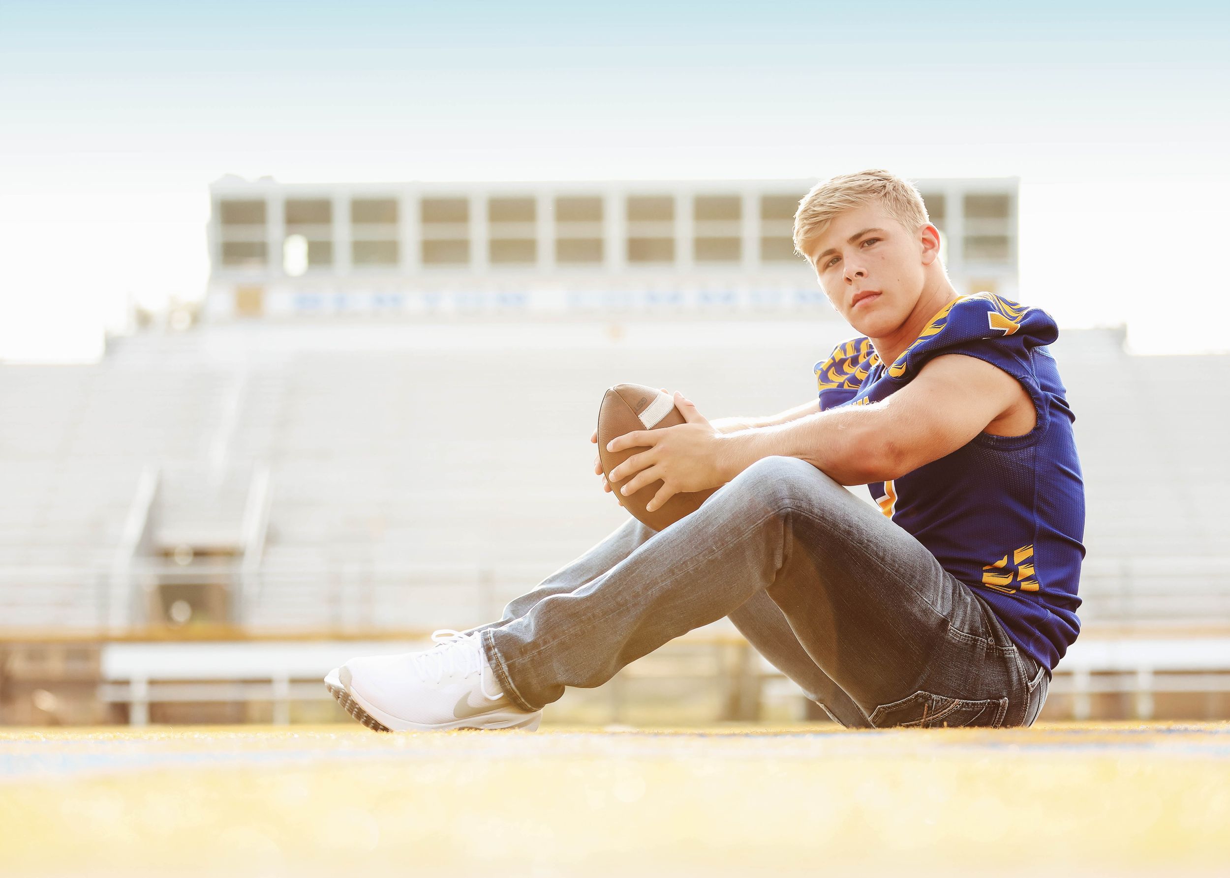Student athlete in blue uniform reclining on stadium field during photo session.