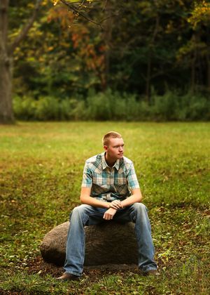 A person in plaid shirt and jeans sits on a fallen log in an autumn forest setting.