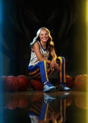 Young female basketball player in blue and gold uniform sitting on court surrounded by basketballs in a professional sports portrait.