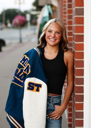 Smiling teenage girl in black crop top and jeans holding a blue and white ST letterman jacket against a brick wall.