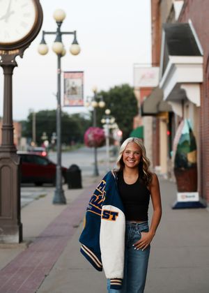Smiling teenage girl holding a varsity jacket walks along a small-town sidewalk near a vintage street clock at dusk.