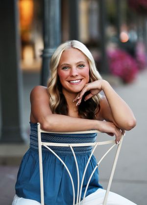 Smiling blonde teenage girl in blue strapless top sitting on white chair outdoors for senior portrait photo.