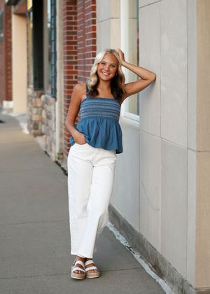Young woman in blue smocked top and white wide-leg pants leans against wall on urban sidewalk.