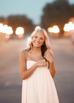 Young woman in white dress smiling outdoors at dusk with bokeh lights in background.