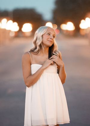 Young woman in white sundress poses outdoors at dusk with bokeh street lights glowing softly in background.