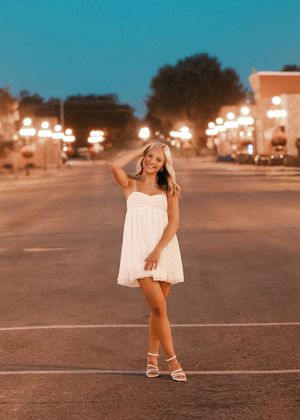 Young woman in white mini dress and heels poses on a street at dusk with glowing streetlights in background.