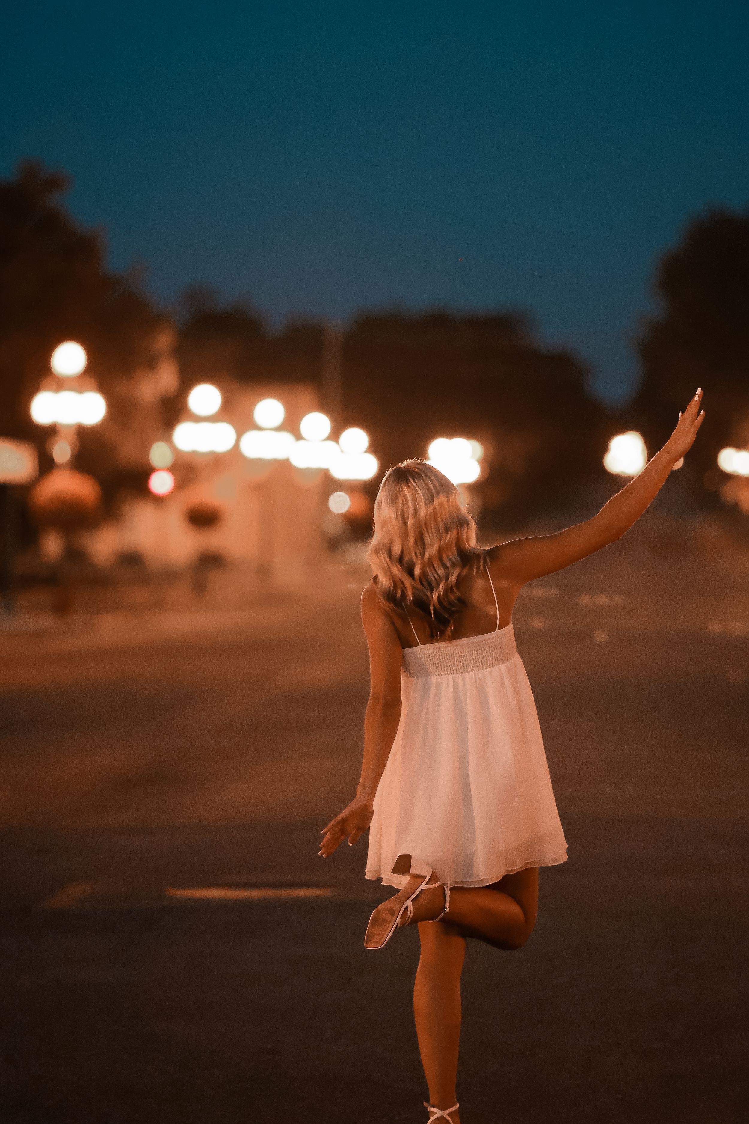 A blonde woman in a white dress poses gracefully on one leg at night, with glowing street lamps illuminating the scene.