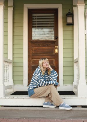 A person in a striped sweater sits casually on a porch step in front of a green house with wooden door.