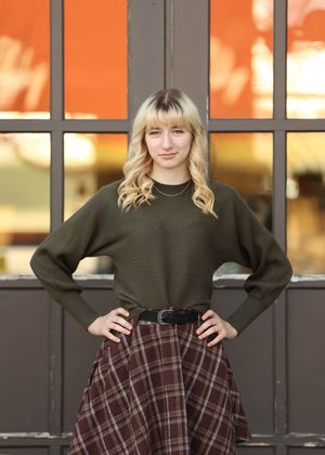 A stylish autumn outfit featuring an olive sweater and plaid skirt against an orange-tinted window background.