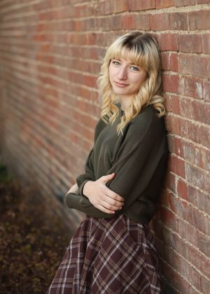 A portrait against a brick wall wearing an olive sweater and plaid skirt with wavy blonde hair.