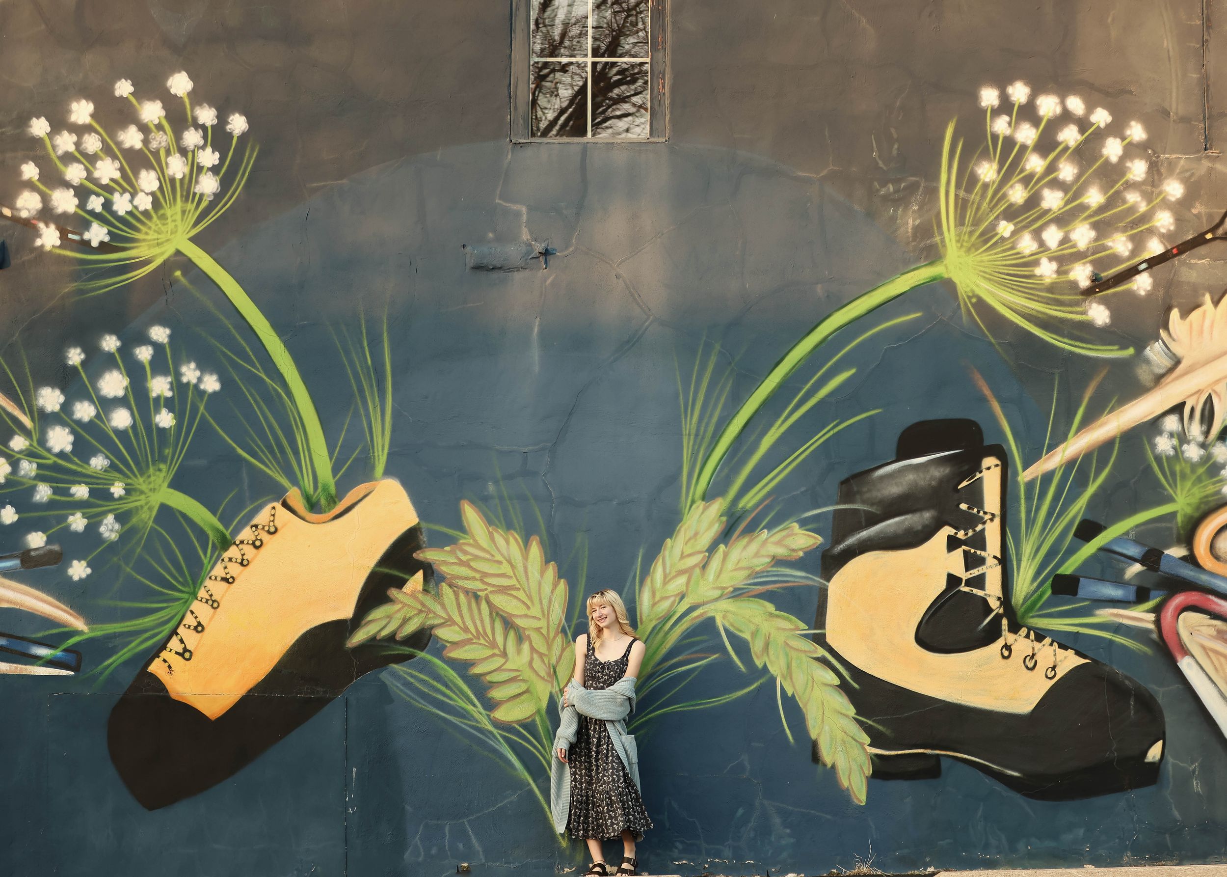 A person poses in front of a vibrant mural featuring giant dandelions and floating shoes.
