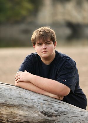 A person in navy clothing leans on driftwood by a waterfront with cliffs in the background.