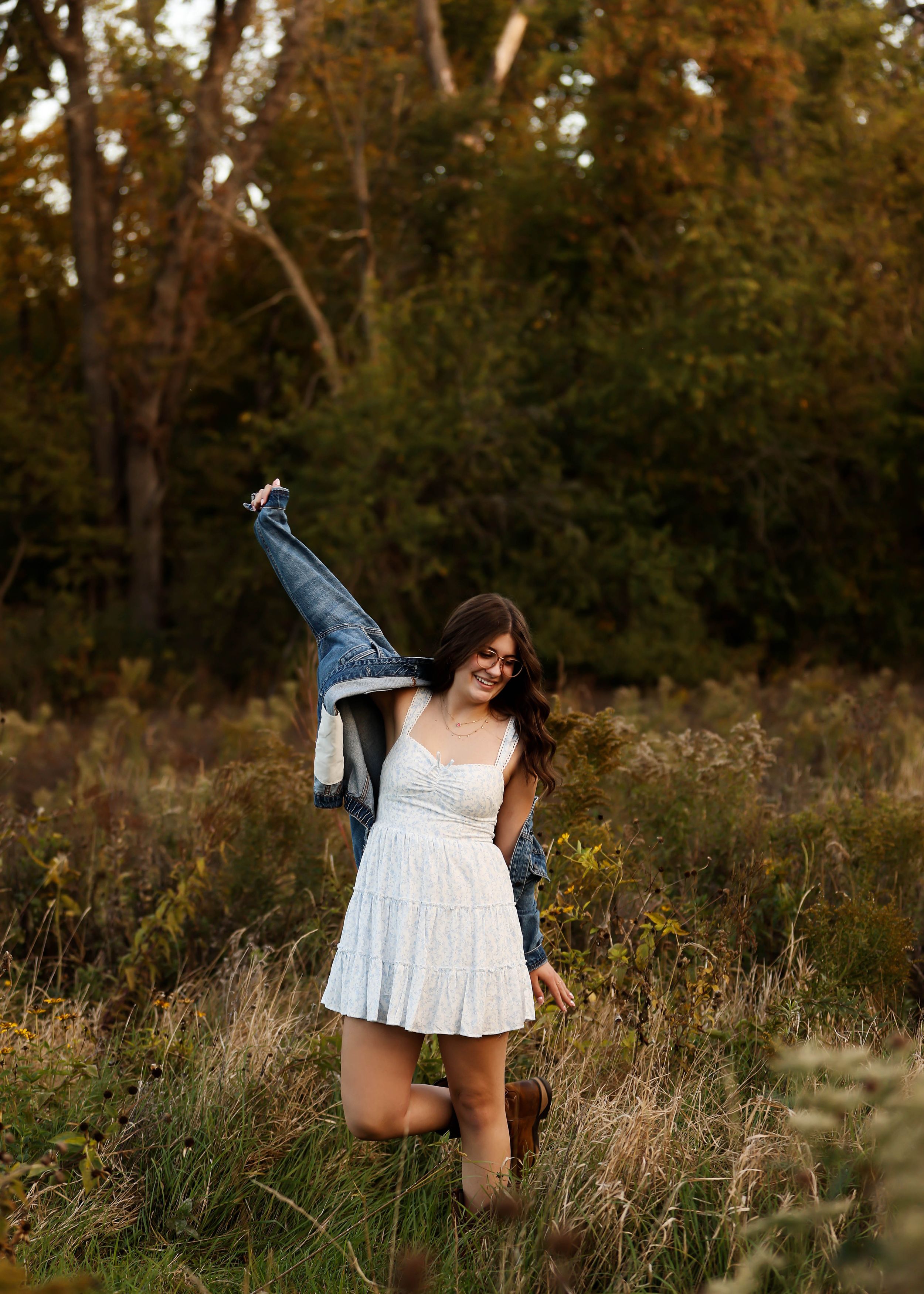 Someone in a white dress twirling with a denim jacket in a field at sunset.