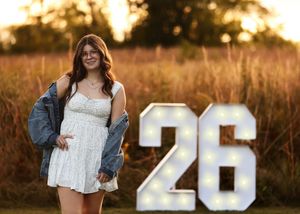 Outdoor portrait in a white dress with denim jacket next to large number 26.