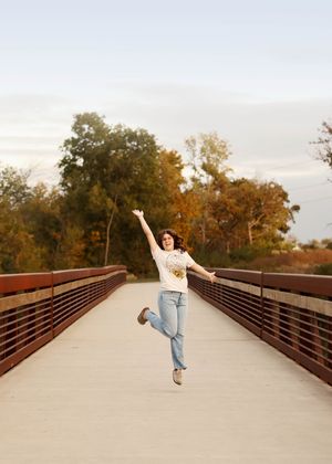 Joyful jumping pose on a wooden bridge with autumn trees in the background.