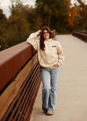 Leaning against bridge railing wearing cream hoodie and jeans in autumn setting.