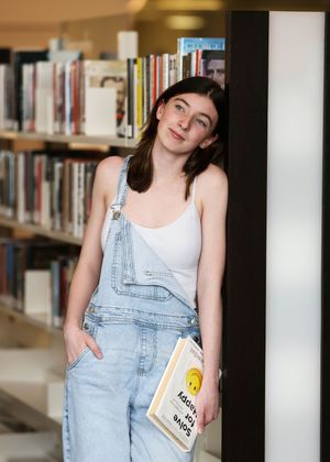 Student in denim overalls holding a book while standing in a library.