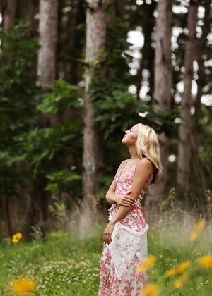 Woman in floral dress standing in wildflower meadow with tall trees, looking up joyfully.