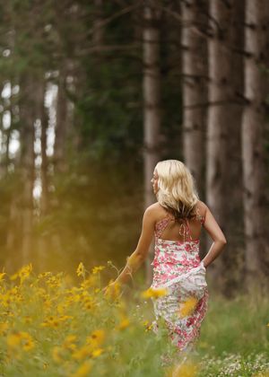 Blonde woman in floral dress walking through wildflower meadow near pine forest on sunny day.