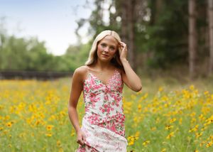 Young blonde woman in floral dress standing in a field of yellow wildflowers with trees in the background.