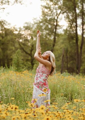 Young blonde woman in floral dress reaching toward sky in vibrant yellow wildflower meadow surrounded by lush green trees.