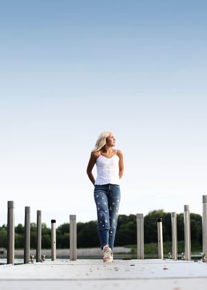 Blonde woman in white tank top and blue jeans walking on a wooden dock near a lake on a clear day.