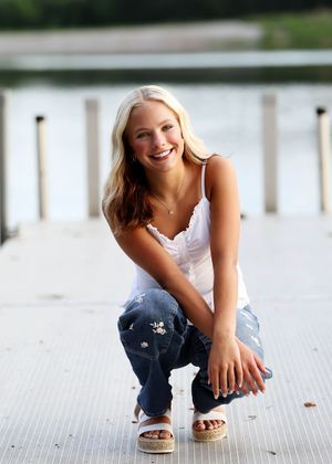 Smiling blonde woman crouching on a wooden dock wearing white lace top, distressed jeans, and wedge sandals.
