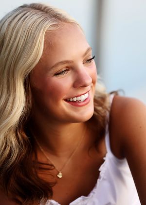 Smiling young blonde woman with a gold necklace wearing a white top, photographed outdoors in warm sunlight.