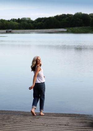 A smiling blonde woman stands barefoot on a wooden dock by a calm lake, wearing jeans and a white tank top.